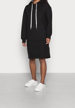 Kobieta ubrana w czarną sukienkę z kapturem z białymi sznureczkami i białymi trampkami, stojąca na jednolitym jasnym tle.