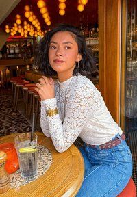 Weißes Spitzenoberteil mit langen Ärmeln und goldenen Blumenmanschetten, hochtaillierte blaue Jeans und runde Ohrringe. Glas Wasser auf einem Holztisch.