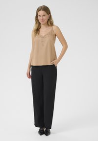 Beige siidist tank-topi, mille kaeluses on pitsdetail, koos kõrge vöökohaga mustade pükstega ja terava ninaga mustade kingadega.