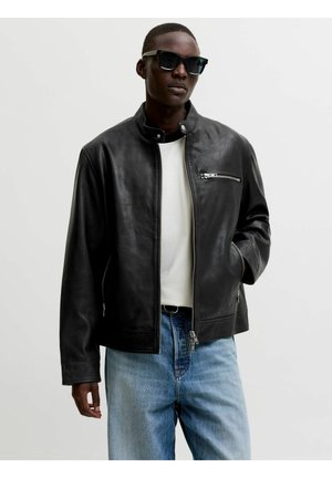 Homme portant une veste en cuir noire, une chemise blanche, un jean bleu et des lunettes de soleil noires, debout avec une main dans la poche de la veste.