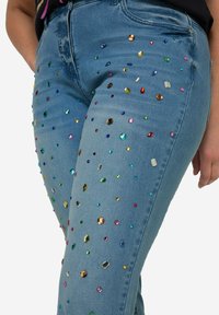 Jeans in denim blu snelli decorati con vari strass multicolori di diverse forme e dimensioni, con un classico design a cinque tasche.