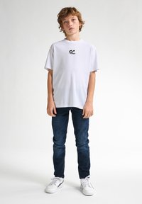 Bílý tričko s krátkým rukávem a malým černým logem na prsou, v kombinaci s tmavě modrými džíny a bílými teniskami. Jednoduché pozadí.