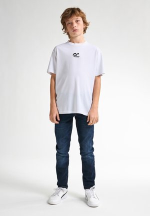 Witte t-shirt met korte mouwen en een klein zwart logo op de borst, gecombineerd met donkerblauwe jeans en witte sneakers. Effen achtergrond.