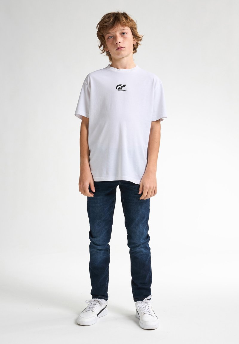 Bílý tričko s krátkým rukávem a malým černým logem na prsou, v kombinaci s tmavě modrými džíny a bílými teniskami. Jednoduché pozadí.