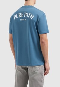 Korte mouw blauwe katoenen t-shirt met witte gebogen tekst "PURE PATH" en "Social Club" op de achterkant. Standaard pasvorm met een ronde hals.