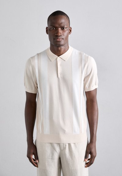 Beige polo shirt met korte mouwen en verticale witte strepen, een knoopsluiting, en een gestructureerde stof. Gecombineerd met lichtgekleurde broeken.