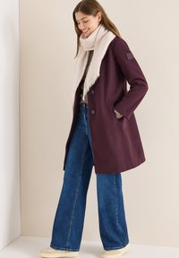 Usměvavá žena v purpurovém kabátě, béžové šále, modrých širokých džínách a krémových teniskách stojící před neutrálním pozadím.
