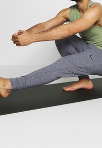 Grå träningstights med en figurnära design och en ljusgrön linne. Personen sitter i en stretchpose på en svart yogamatta.