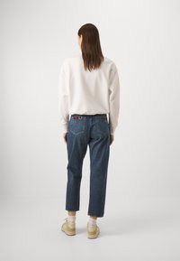 Vit sweatshirt, blå denimjeans med en något lös passform och beiga skor. Synlig flerfärgad söm på jeansens linning.
