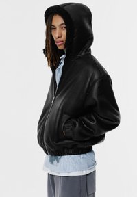 Svart bomberjacka med en mjuk, pälsfodrad huva, dragkedja framtill, ribbade muddar, avslappnad passform, bärs över en ljusblå randig skjorta.