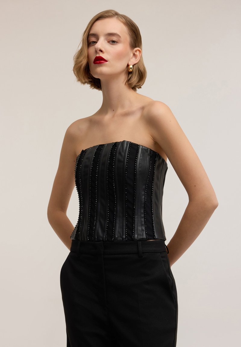 Corsetto nero senza spalline con pieghe verticali, decorato con piccole perline, abbinato a pantaloni neri a vita alta.