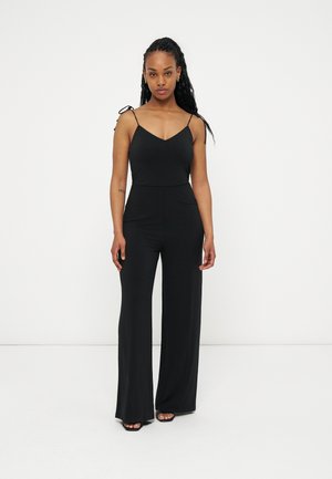 Žena stojí čelem vpřed, oblečená v černém overal bez rukávů s širokými nohavicemi a ramenními pásky na zavazování, na jednobílém pozadí.