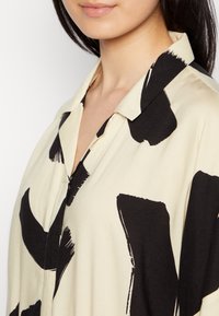 Blusa beige con cuello que presenta patrones abstractos en pinceladas negras. Tejido suave con un corte holgado y cierre de botones.