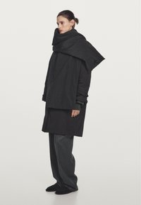 Cappotto nero oversize con sciarpa a strati, caratterizzato da un design testurizzato e una vestibilità ampia, abbinato a pantaloni larghi scuri e scarpe nere.