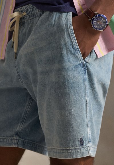 Άτομο που φοράει ανοιχτόχρωμα τζιν σορτς με σούρα στη μέση και χέρι στην τσέπη, πολύχρωμο ριγέ πουκάμισο και ρολόι με καφέ δερμάτινο λουράκι.