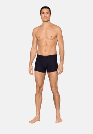 Shirtless man with long dark hair tied back wearing black boxer briefs, standing barefoot against a plain light background.