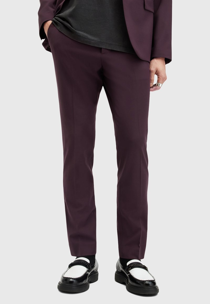 Pantalons ajustés bordeaux avec une finition en tissu lisse, coupe slim et détails de pli minimal, associés à des chaussures noires et blanches.