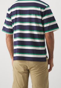 Mężczyzna w granatowej, białej i zielonej, poziomo paskowanej koszulce z krótkim rękawem oraz khaki szortach, widoczny od tyłu na jednolitym tle.