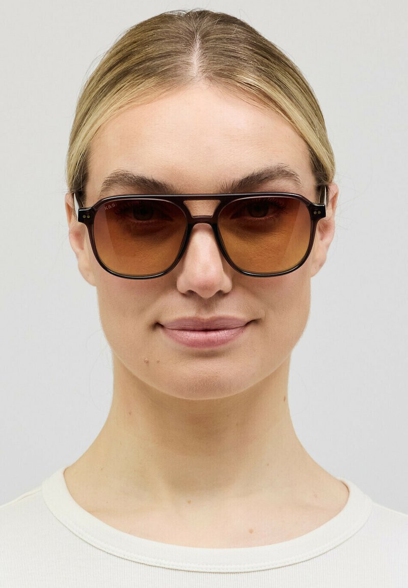 Vrouw met licht haar dat naar achteren is gebonden, draagt grote bruingetinte rechthoekige zonnebril en een wit T-shirt met ronde hals, tegen een effen achtergrond.