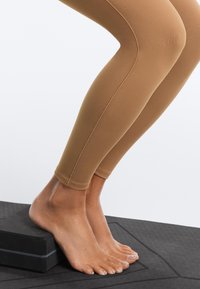 Pieds nus appuyant les talons sur un bloc de yoga noir posé sur un tapis texturé, personne portant des leggings beiges, démontrant un étirement du mollet.