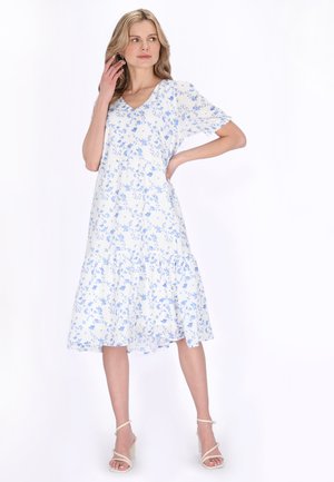 Kvinde i en hvid midikjole med blåt blomsterprint, puffede ærmer og lagdelt skørt, iført beige hæle med stropper, poserer med den ene hånd på hoften.