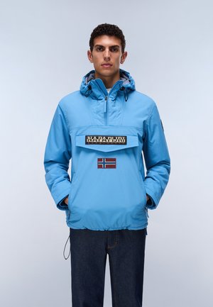 Junger Mann, der eine hellblaue Kapuzenjacke mit einer Fronttasche und Logoaufnäher trägt, steht mit den Händen in den Taschen vor einem schlichten Hintergrund.