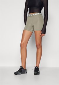 Nike Pro kratke hlače u maslinasto zelenoj boji s sivim pojasom. Materijal izgleda rastezljivo. Nosene s crnim sportskim cipelama i prozirnom crnom dugom majicom.