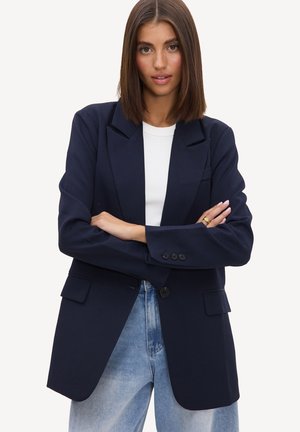 Femme aux cheveux bruns raides portant un blazer bleu marine foncé sur un haut blanc et un jean bleu, debout les bras croisés sur fond blanc.