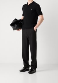 Zwart poloshirt met korte mouwen en logo, gecombineerd met zwarte broek en glanzende zwarte schoenen. Houdt een zwarte leren tote bag vast.