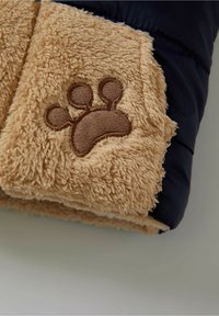 Tanfarbener, flauschiger Stoff mit besticktem Pfotenabdruck auf einem warmen, gesteppeten navyblauen Abschnitt. Weicher Textil, kompaktes Design, geeignet für Haustiere.