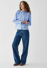 Camicia button-up a righe di un blu chiaro con dettagli arricciati, abbinata a jeans blu scuro dal fit ampio e scarpe marroni.