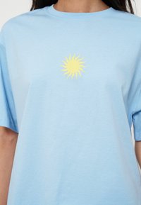 T-shirt bleu clair avec un petit motif de soleil jaune au centre de la poitrine, porté par une personne aux longs cheveux foncés.