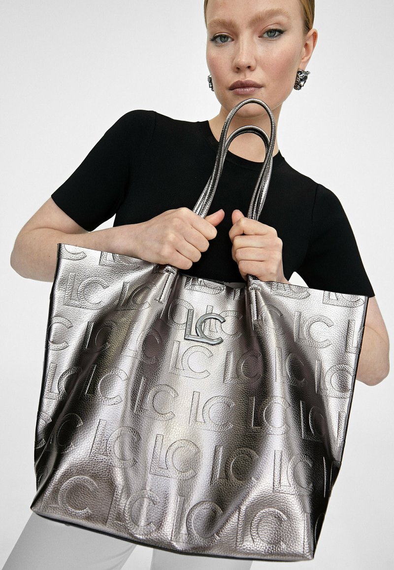 Kobieta w czarnej bluzce trzyma dużą metaliczną srebrną torbę na ramię z wypukłym wzorem logo "LC" i podwójnymi uchwytami.
