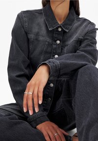 Veste en denim noir avec col, boutons argentés et deux poches latérales. Le tissu présente une texture délavée, associée à un jean noir assorti.