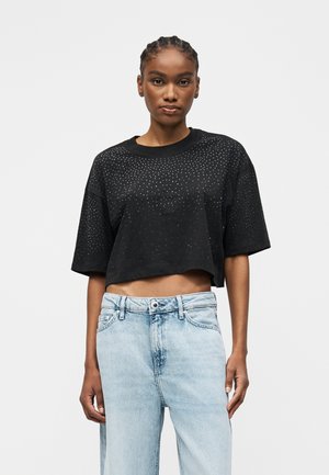 Mujer con cabello trenzado que lleva una camiseta negra corta con pequeños puntos blancos y vaqueros de tiro alto azul claro sobre fondo blanco.