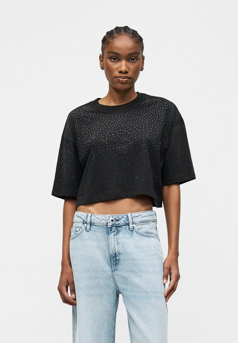 Mujer con cabello trenzado que lleva una camiseta negra corta con pequeños puntos blancos y vaqueros de tiro alto azul claro sobre fondo blanco.