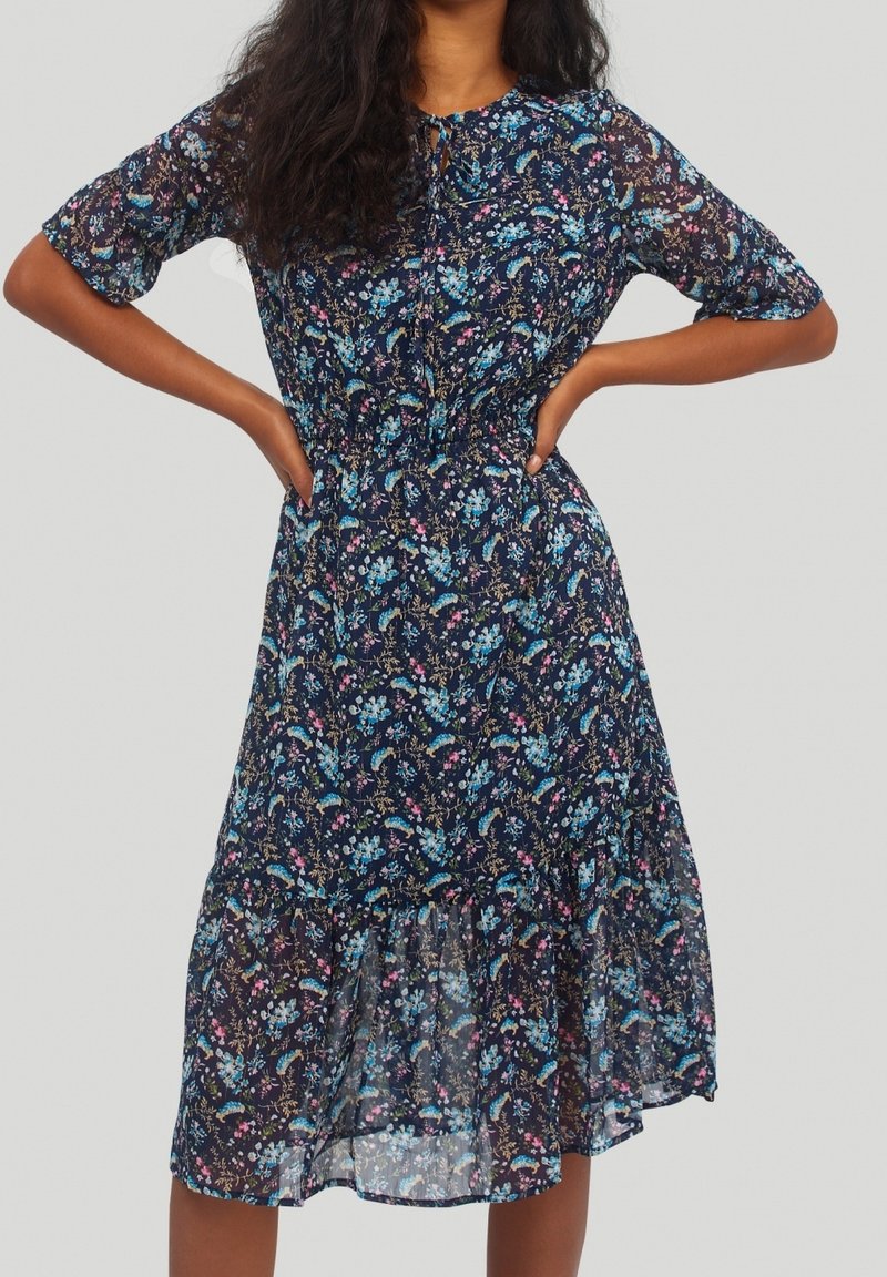Granatowa sukienka w kwiaty z krótkimi rękawami, zebranym w pasie i wielowarstwowym falbanym dołem. Cechuje się mieszanką niebieskich i różowych wzorów kwiatowych.