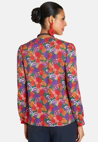 A woman with dark hair in a bun wears a vibrant floral blouse featuring red, purple, and orange flowers, paired with red heart earrings.