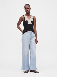 Woman wearing a black sleeveless top with white lace detail, light blue wide-leg jeans, and black sandals, standing with one hand on hip.