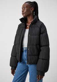 Jachetă puffer neagră cu o suprafață texturată, siluetă supradimensionată, guler înalt și închidere cu nasturi prin apăsare. Purtați peste un top gri și blugi bleu.
