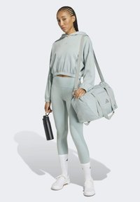 Lysegrøn atletisk sweatshirt, matchende leggings, hvide sneakers, holder en sort vandflaske og en lysegrå træningstaske med detaljer.