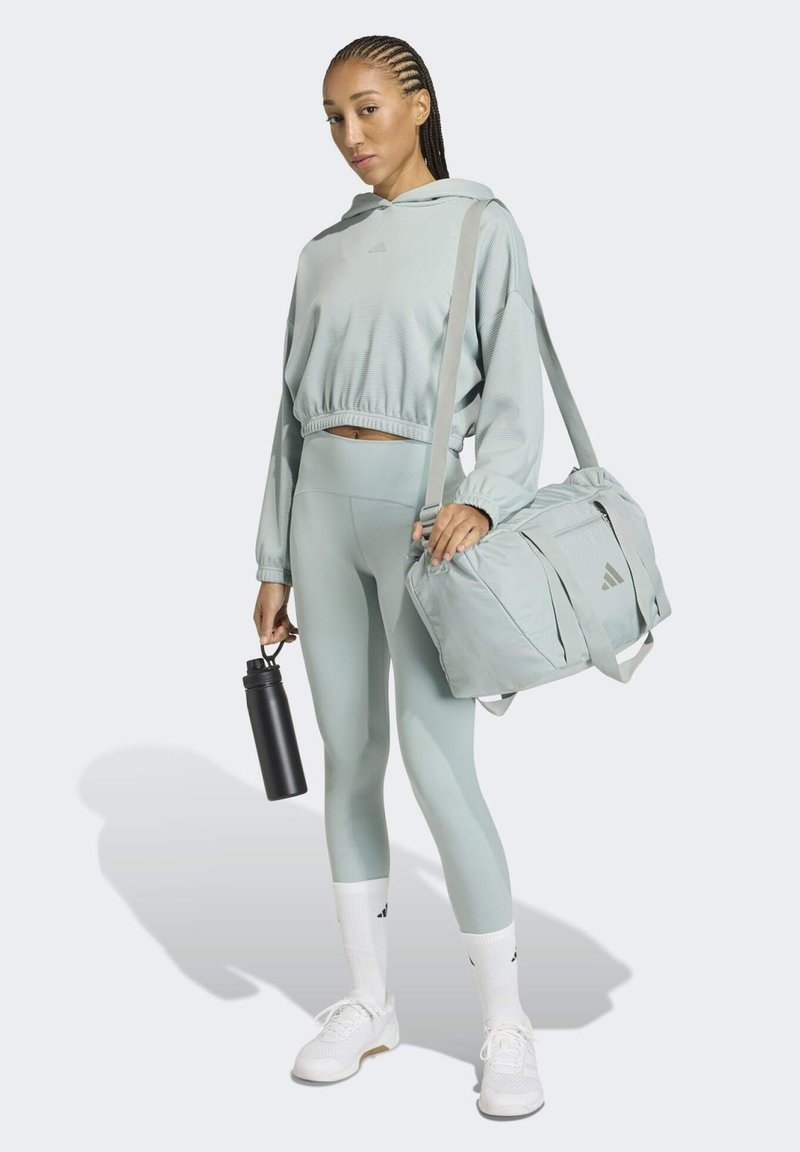 Lysegrøn atletisk sweatshirt, matchende leggings, hvide sneakers, holder en sort vandflaske og en lysegrå træningstaske med detaljer.