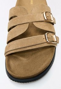 Hnědá semišová sandála s dvěma širokými překříženými pásky se zlatými přezkami a texturovanou stélkou, umístěná na černé gumové podrážce.
