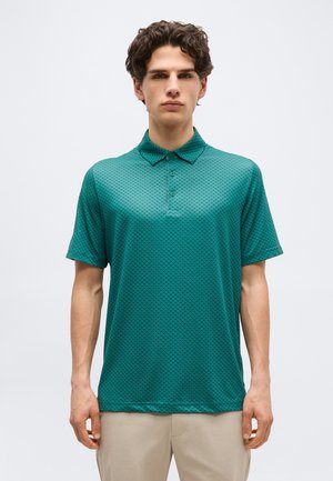 Joven con cabello rizado oscuro que lleva una camisa tipo polo de manga corta con estampado verde y pantalones beige, sobre un fondo sencillo.