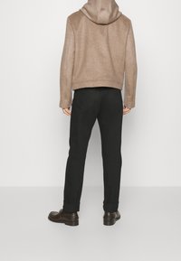 Person i en ljusbrun huva jacka, svarta byxor med uppvikt fåll och mörkbruna läderskor, stående med ansiktet mot en plain vit vägg.