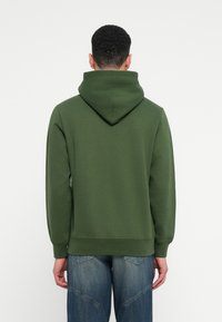 Zaļa kapucē ar lielu, salocītu kapuci, ribotām piedurknēm un apakšmali, izgatavota no mīksta, teksturēta auduma, valkāta ar ziliem džinsiem.