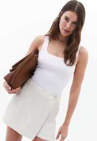 White ribbed tank top, cream wrap skirt with a side button, holding a woven brown handbag. Smooth texture and casual design.