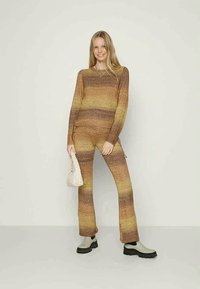 Conjunto de malha em duas peças em tons de castanho e dourado com textura canelada. As calças apresentam aberturas laterais; a modelo segura uma pequena mala branca.