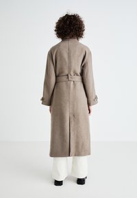 Casaco longo de lã bege com cintura cingida, mangas oversized e punhos com botões; apresenta uma fenda central nas costas e um design de gola plana.