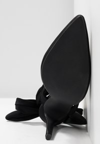 Chaussure noire à talon haut avec semelle pointue reposant sur le côté, avec un talon stiletto fin et une tige en tissu doux.